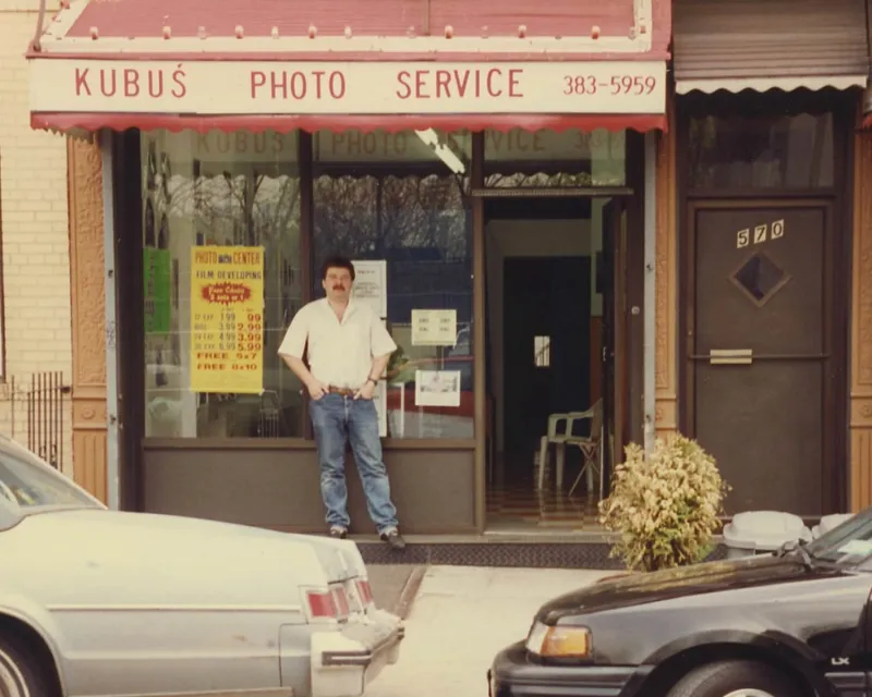 A man with dark hair and a mustache wearing a white shirt and blue jeans standing in front of a photo store named 'Kubuś Photo Service.' There are older cars parked in front of the store, and the store has a large window with signs and posters.