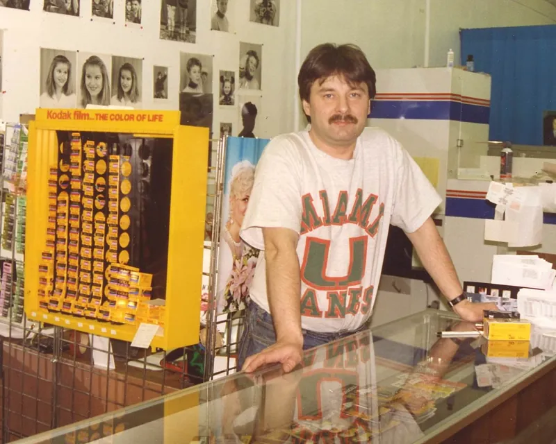 A man with a mustache wearing a Miami Hurricanes T-shirt standing behind a glass counter at a store, with a vending machine of Kodak film and photographs of women on the wall in the background.
