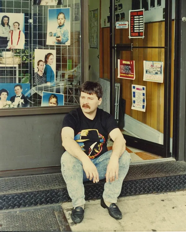 A man with dark hair and a mustache sitting on a step outside a storefront with photographs in the window behind him. The man is wearing a black t-shirt with a colorful design, light-colored acid wash jeans, and black shoes. The storefront has a glass door and window displaying pictures of children and adults.