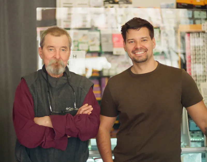 Andrzej and Jakub Redziniak smiling at the camera. The man on the left has a beard, is wearing a vest, and glasses hanging around his neck. The man on the right has short dark hair, a goatee, and is wearing a brown t-shirt.