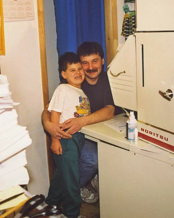 A man with a mustache and a young boy with a big smile, standing together at a desk in an office space, hugging and posing for the photo. The office has stacks of papers, a filing cabinet, and a blue curtain in the background.
